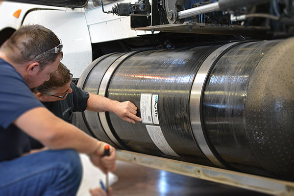 Two vehicle inspectors squatting down to check the label on a CNG cylinder.